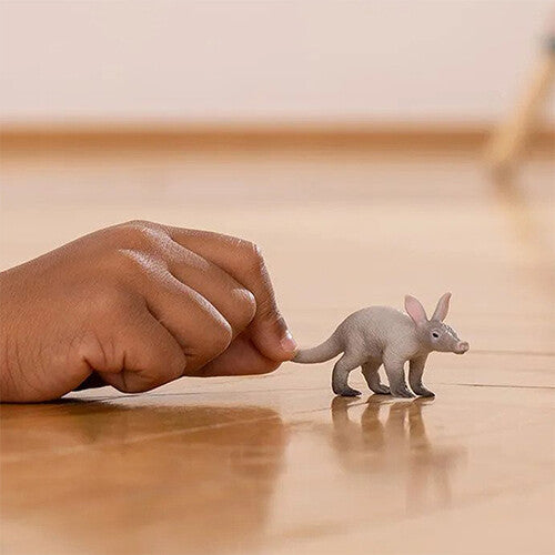 Een klein aardvarken dierenfiguurtje op een houten vloer naast een kinderhand, voor schaal.