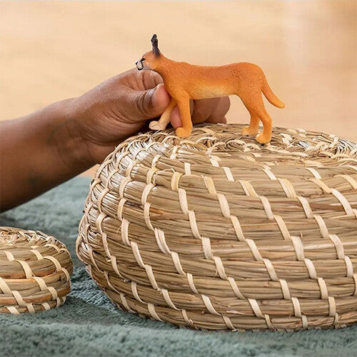 Een hand plaatst het Schleich plastic dierenfiguurtje van een caracal op een gevlochten mand.