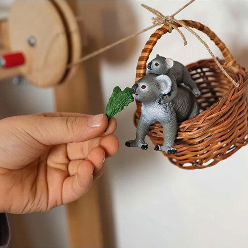 Schleich dierenfiguurtje van een koalabeer met jong in een rieten mandje, waaraan een hand een groen blad aanbiedt.