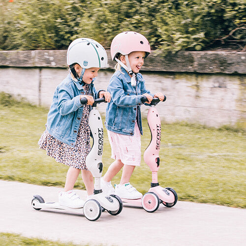 Twee lachende meisjes met helmen rijden samen op een beige en een roze Scoot & Ride 2-in-1 step.