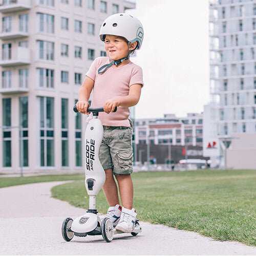 Een lachende jongen met helm staat op een beige Scoot & Ride 2-in-1 step op een pad buiten.