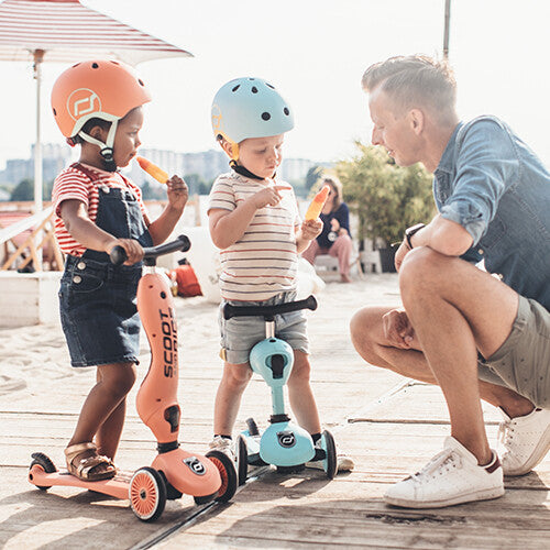 Een volwassen man knielt bij twee jonge kinderen met helmen op hun Scoot & Ride Highwaykick 1 2-in-1 loopfiets en step.