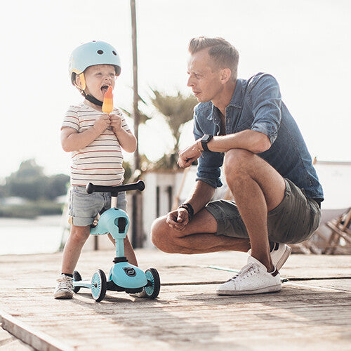 Een volwassen man knielt bij een jong kind met een helm op een blauwe Scoot & Ride Highwaykick 1 2-in-1 loopfiets en step.