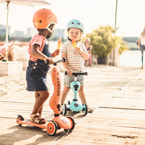 Twee jonge kinderen met helmen op een houten vlonder, rijdend op een oranje en een blauwe Scoot & Ride Highwaykick 1 2-in-1 loopfiets en step.