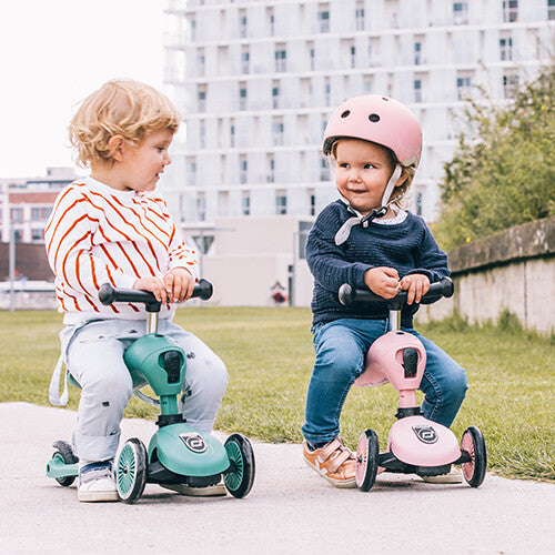 Twee lachende kinderen met helmen op zitten op een groene en een roze Scoot & Ride Highwaykick 1 loopfiets.