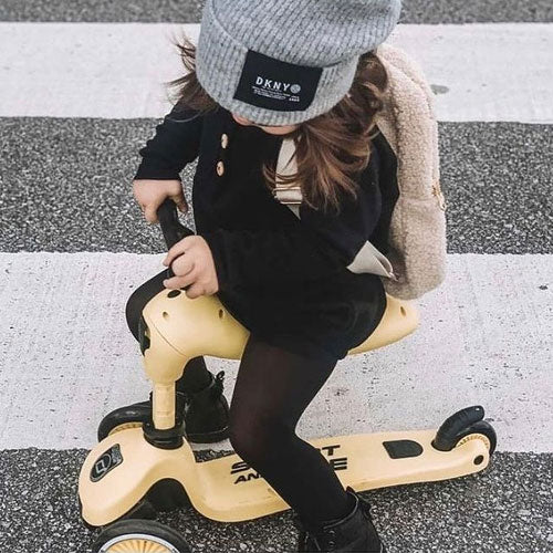 Een kind zit op een gele Scoot & Ride Highwaykick 1 2-in-1 loopfiets in loopfietsmodus op een zebrapad.