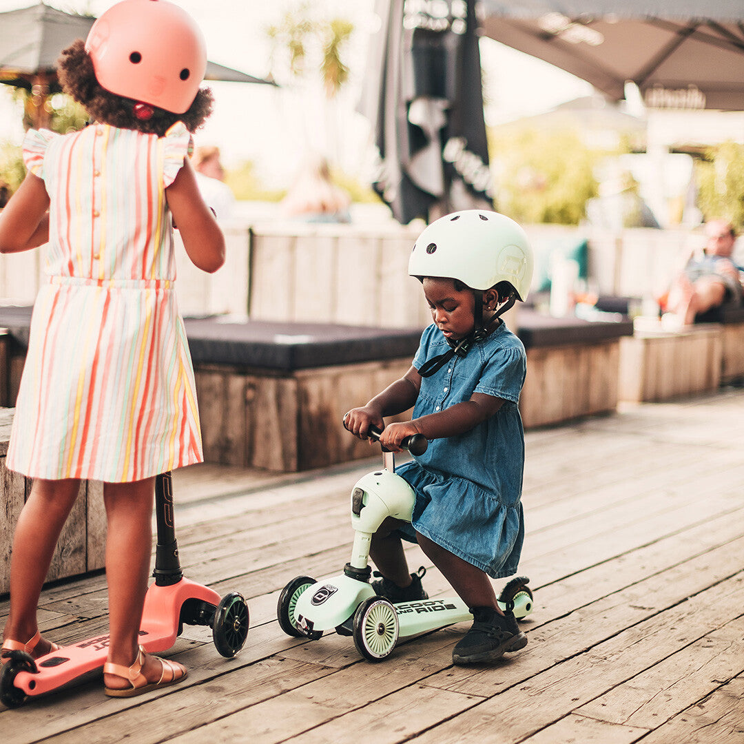 Twee kinderen op een houten terras, één zit op een lichtgroene Scoot & Ride Highwaykick 1 loopfiets en de ander staat bij een roze step.