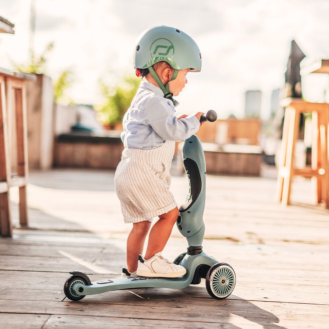 Een kind met helm staat op een groene Scoot & Ride Highwaykick 1 step op een houten terras.