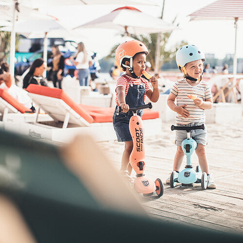 Twee jonge kinderen met helmen eten ijsjes, staand bij hun Scoot & Ride Highwaykick 1 2-in-1 loopfietsen en steps op een houten vlonder.