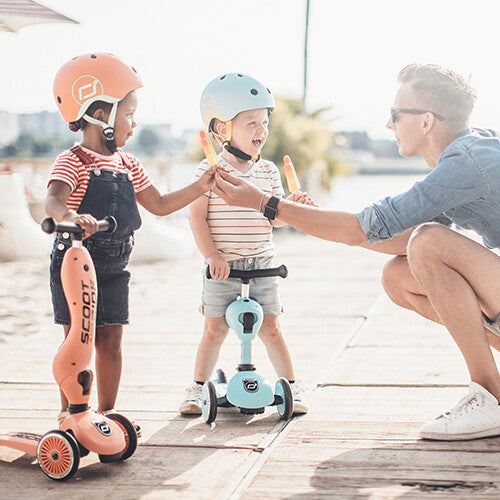 Een man geeft ijsjes aan twee jonge kinderen met helmen, staand bij hun Scoot & Ride Highwaykick 1 2-in-1 loopfietsen en steps op een houten vlonder.