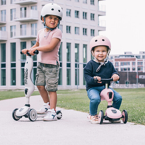Twee jonge kinderen met helmen buiten, één op een witte Scoot & Ride Highwaykick 1 step en de ander op een roze loopfiets.