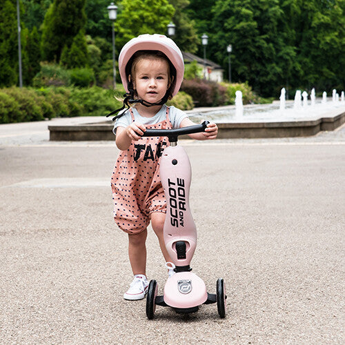 Een jong kind met roze helm staat met een roze Scoot & Ride Highwaykick 1 step in een park.