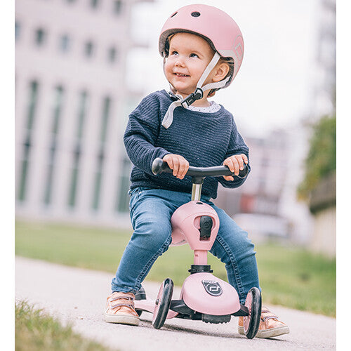 Een lachend kind met roze helm zit op een roze Scoot & Ride Highwaykick 1 loopfiets.