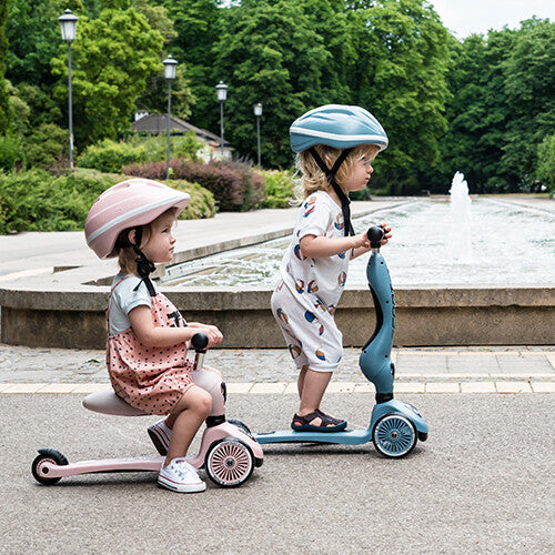 Twee kinderen in een park, één op een roze Scoot & Ride Highwaykick 1 loopfiets en één op een blauwe step.