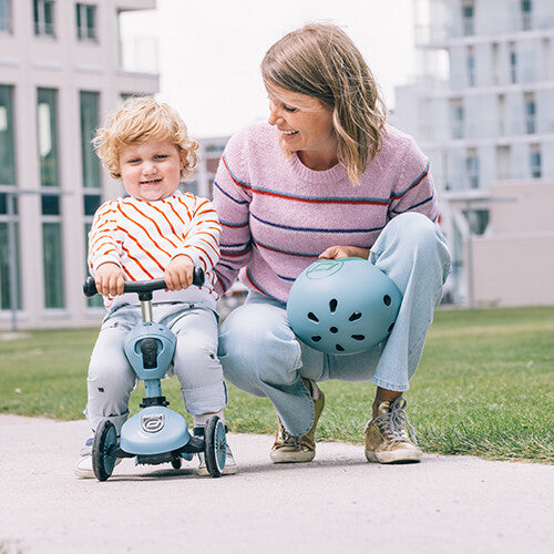 Een peuter zit op een staalblauwe Scoot & Ride Highwaykick 1 loopfiets naast een lachende vrouw met een helm.