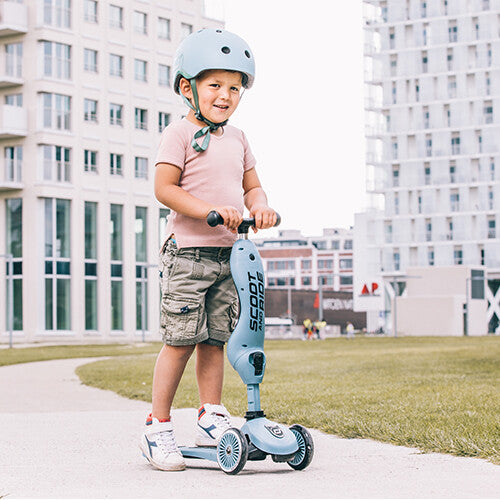 Een lachende jongen met helm rijdt op een staalblauwe Scoot & Ride Highwaykick 1 step buiten.