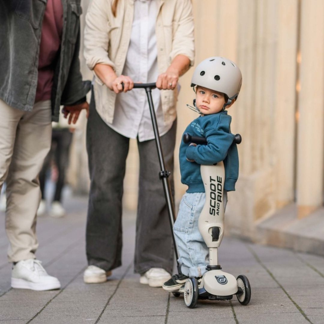 Een kind met helm staat op een beige Scoot & Ride 2-in-1 step, met volwassenen op de achtergrond.