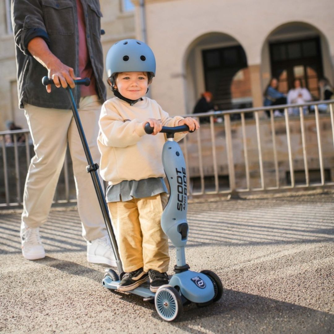 Een kind met helm rijdt op de blauwe Scoot & Ride 2-in-1 step, geduwd door een volwassene.