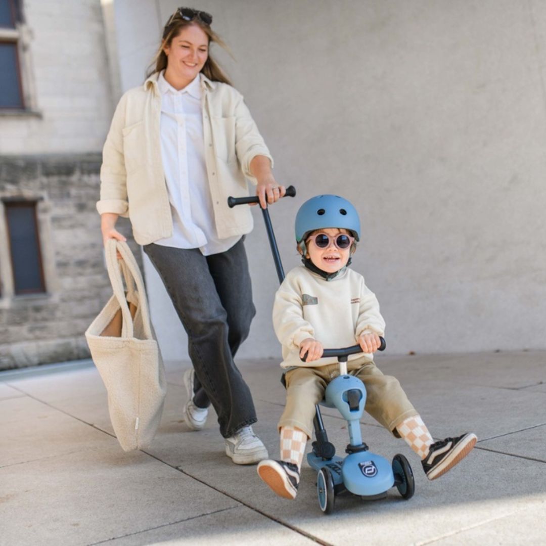 Een kind met helm en zonnebril rijdt op de blauwe Scoot & Ride 2-in-1 loopfiets, geduwd door een volwassene.