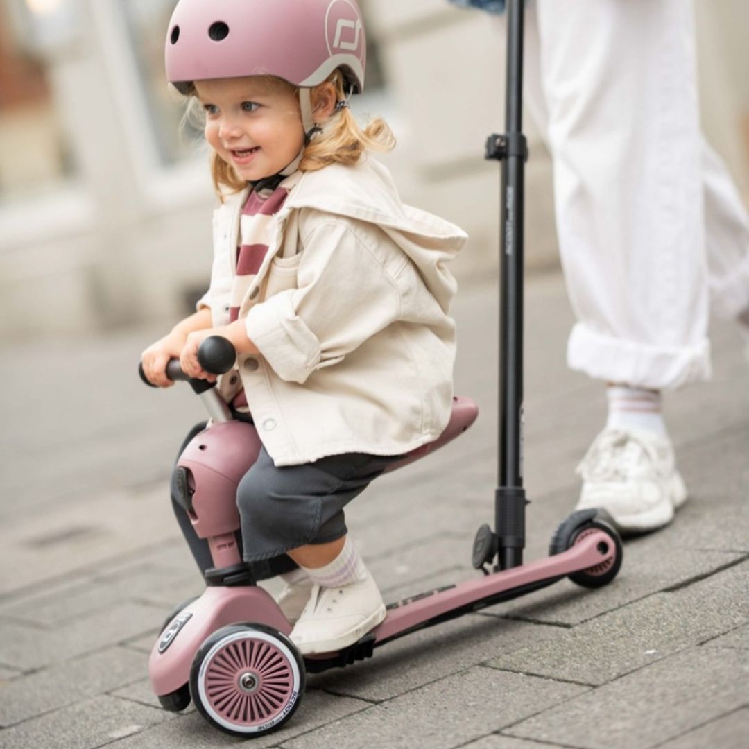 Een lachend kind met roze helm rijdt op de roze Scoot & Ride 2-in-1 loopfiets, geduwd door een volwassene.