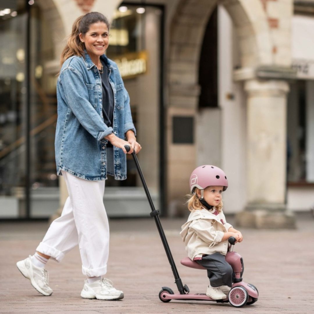 Een vrouw duwt een kind met roze helm op de roze Scoot & Ride 2-in-1 loopfiets over een plein.