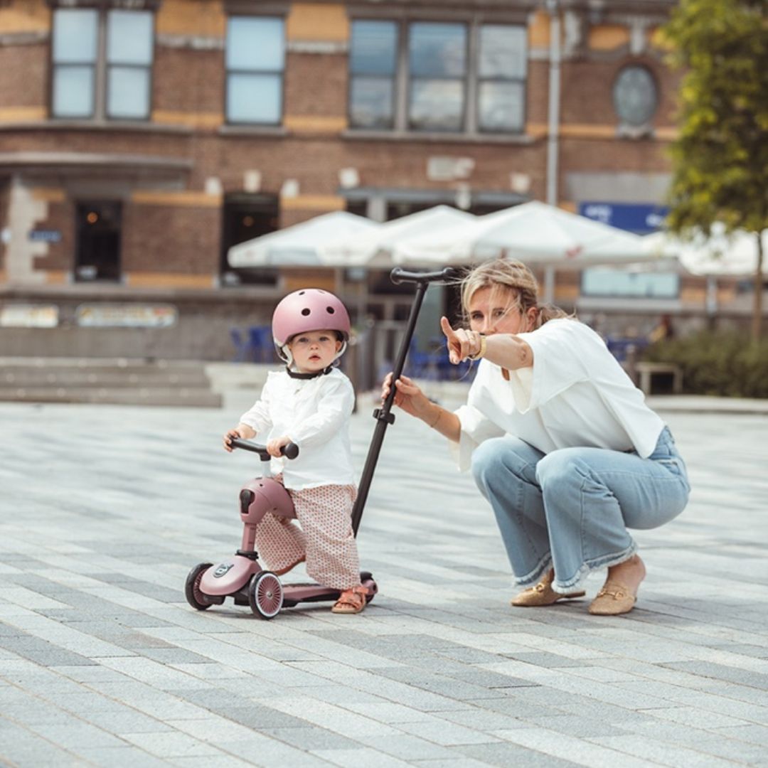Een kind met roze helm zit op de roze Scoot & Ride 2-in-1 loopfiets, terwijl een volwassene ernaast knielt en wijst.