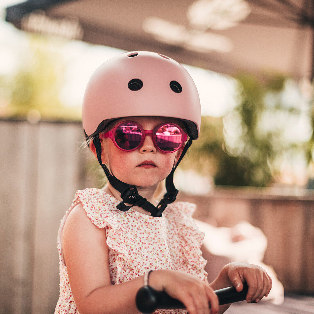 Een kind met een roze kinderhelm en roze zonnebril op, kijkend naar de camera.