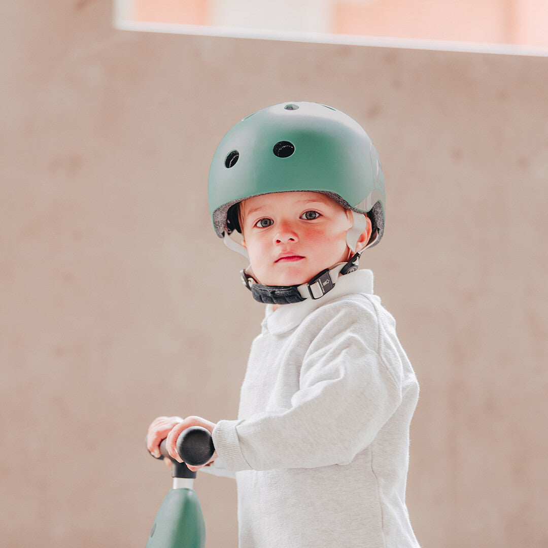 Een kind met een groene Scoot & Ride kinderhelm op, staand bij een loopfiets.