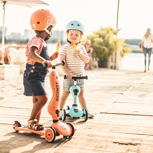 Twee kinderen met kinderhelmen op Scoot & Ride scooters, buiten spelend met een ijsje.