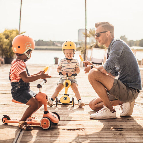 Twee kinderen met kinderhelmen op Scoot & Ride scooters en een volwassene die naast hen knielt.