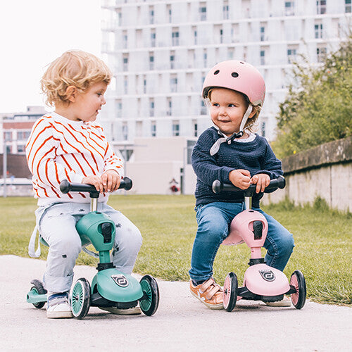 Twee kinderen buiten op loopfietsjes, waarvan één een roze Scoot & Ride kinderhelm draagt.