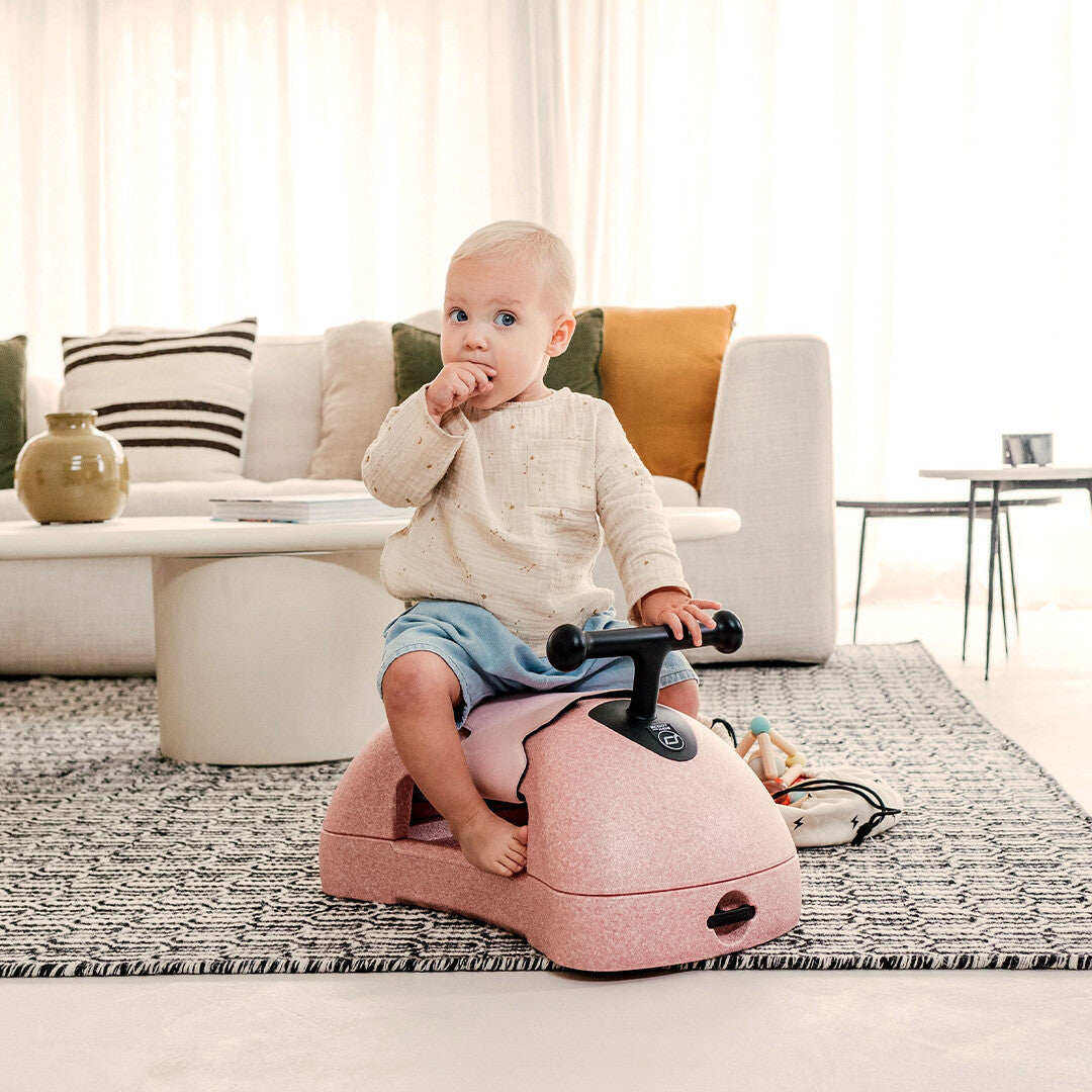 Een baby zit op de Scoot & Ride roze loopwagen van schuim in een woonkamer.