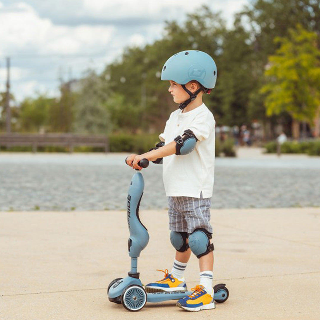 Een kind met een blauwe Scoot & Ride helm, elleboog- en kniebeschermers staat op een blauwe scooter.