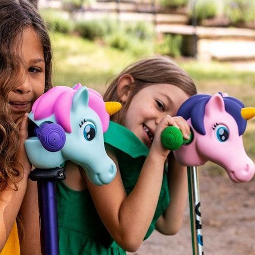 Twee lachende kinderen met scootaheadz kinderstep accessoires, een mintkleurig en een roze eenhoornhoofd.