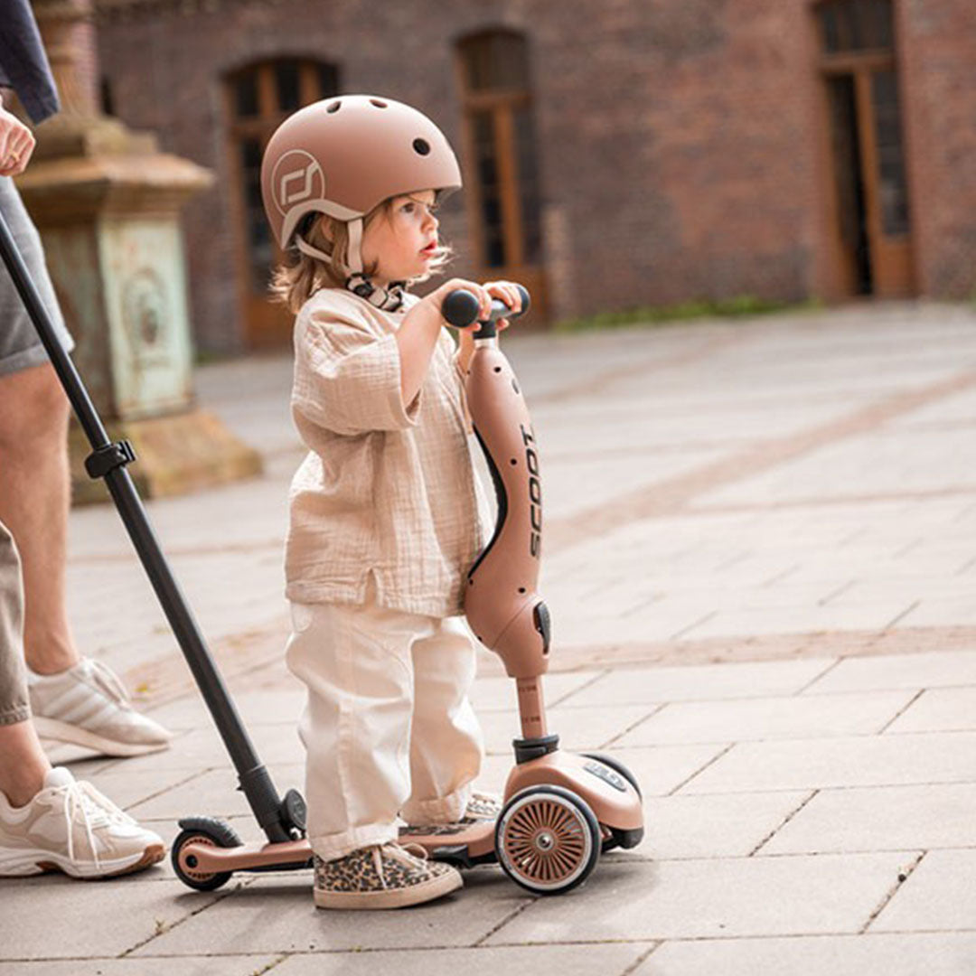 Een kind met helm staat op een Scoot & Ride 2-in-1 step in mocha mousse kleur, voortgeduwd door een volwassene.