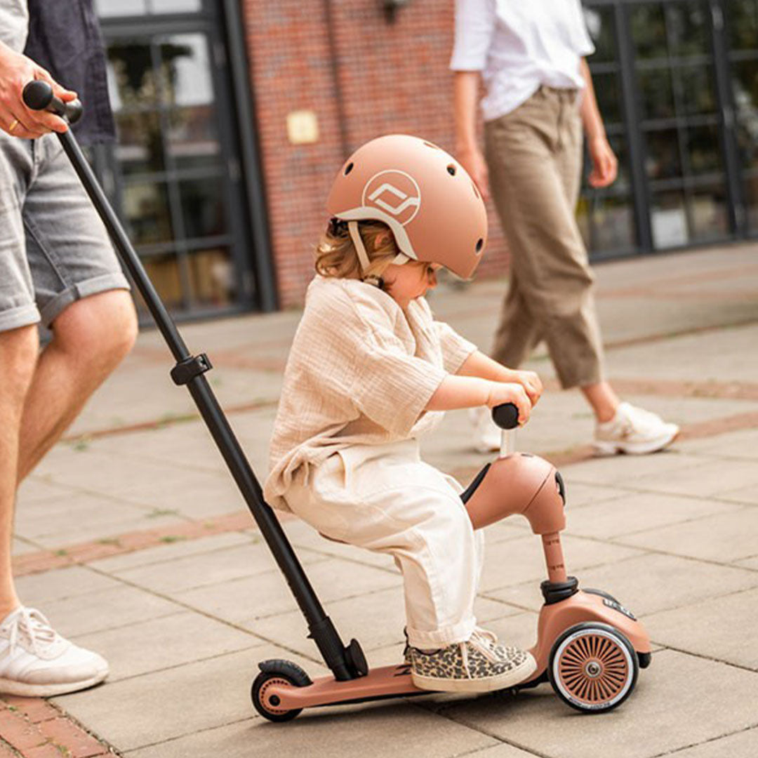 Een kind met helm zit op een Scoot & Ride 2-in-1 loopfiets in mocha mousse kleur, voortgeduwd door een volwassene.
