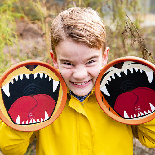 Een jongen lacht en houdt twee oranje vangschijven met tijger/leeuwenmonden voor zijn gezicht.