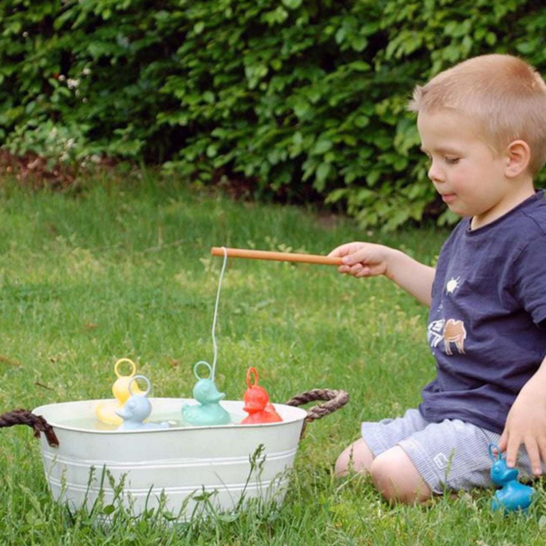 Een jongen vist met een houten hengel op gekleurde eendjes in een witte teil op het gras.