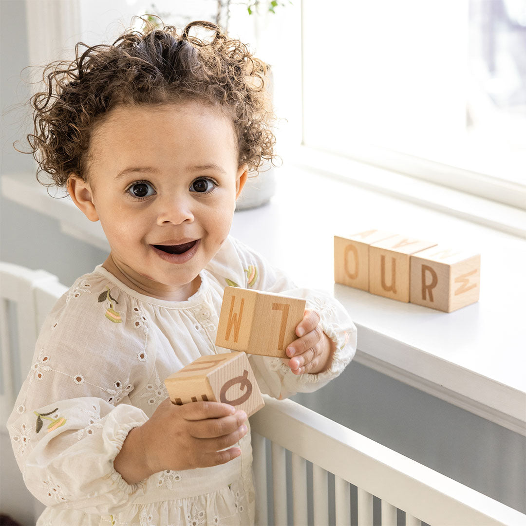 Een lachende baby speelt met Sebra houten alfabetblokken bij een raam.