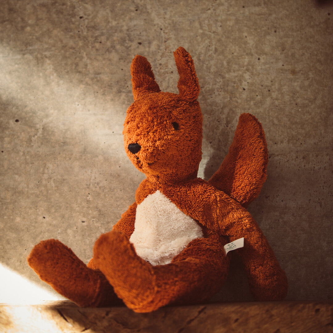 De bruine Senger pluchen knuffeleekhoorn zit tegen een grijze muur.