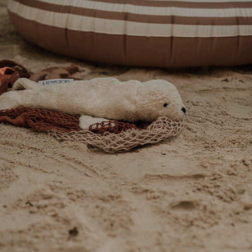 senger witte pluchen zeehond warmteknuffel liggend op een bruin net in het zand.