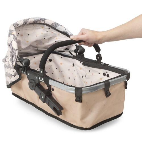 Detail van de Skrållan poppenwagen reiswieg in beige met abstract patroon, vastgehouden door een hand.