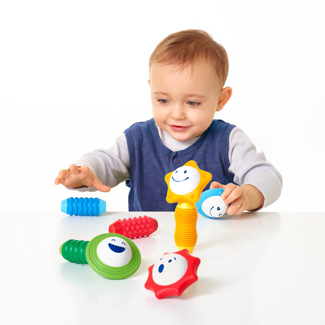 Een baby kijkt aandachtig naar de Smartmax My First Sounds & Senses rammelaars op een witte tafel.