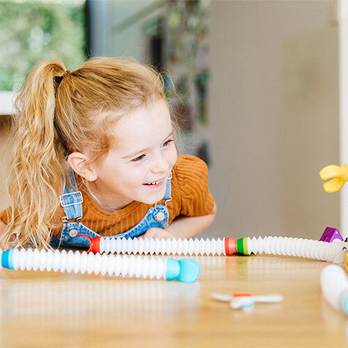 Een lachend meisje kijkt naar de flexibele onderdelen van de Smartmax Roboflex bouwset op een houten tafel.