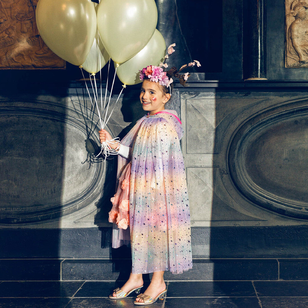 Een meisje draagt de souza for kids veelkleurige kinder cape met sterren en capuchon, terwijl ze ballonnen vasthoudt in een klassieke ruimte.