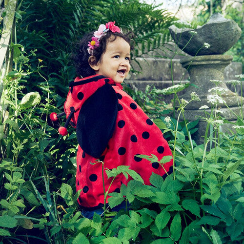 Een lachend kind draagt de Souza for Kids lieveheersbeestje cape in een groene tuin.