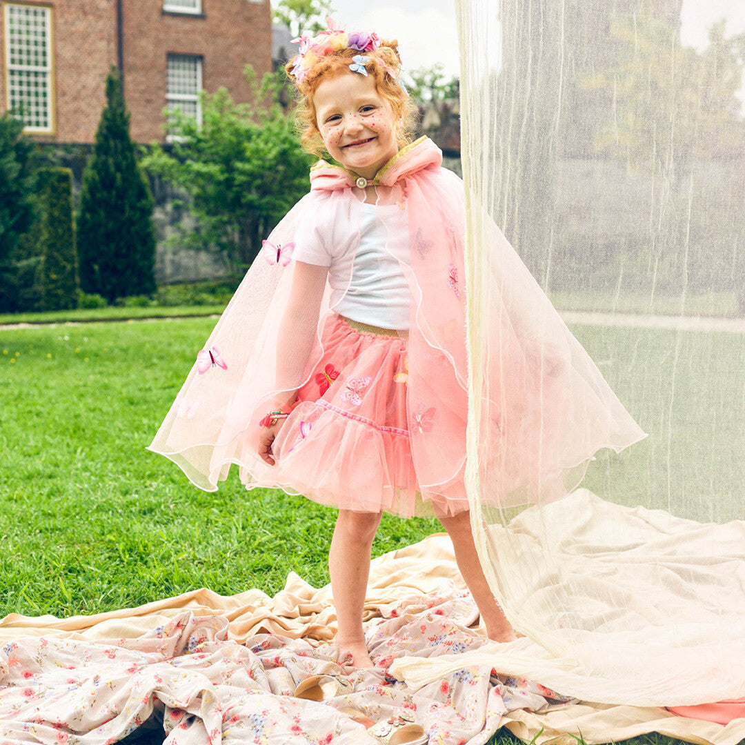Een lachend meisje draagt de roze kinder cape met vlinders en een roze rok in een tuin.