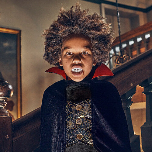 Een kind met krullend haar draagt een Souza for Kids Dracula cape en vampiertanden.