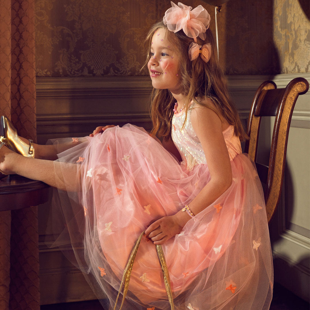Een meisje draagt de roze feestjurk met vlinders, zittend op een stoel met haar voeten op tafel.