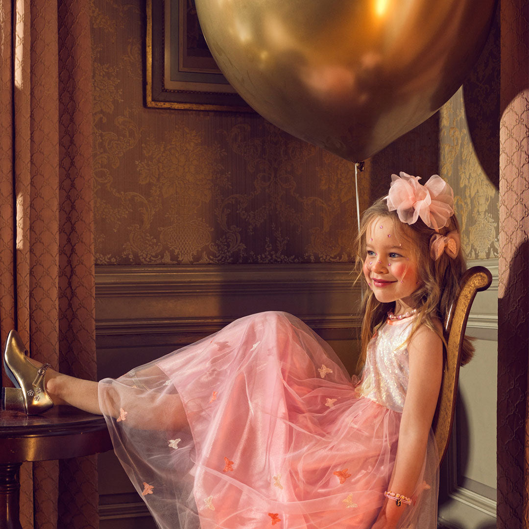 Een lachend meisje draagt de roze kinderfeestjurk met vlinders, met een gouden ballon op de achtergrond.